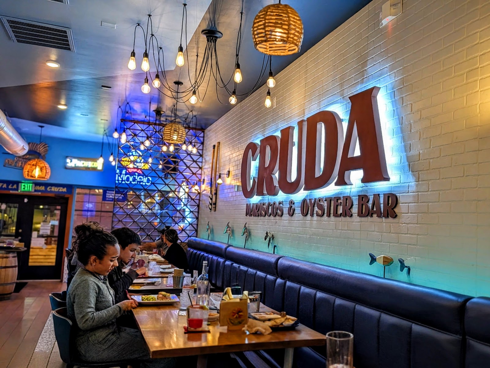 Guests eating along the banquette under Edison-bulb clusters and neon beer signs in the CRUDA dining room.