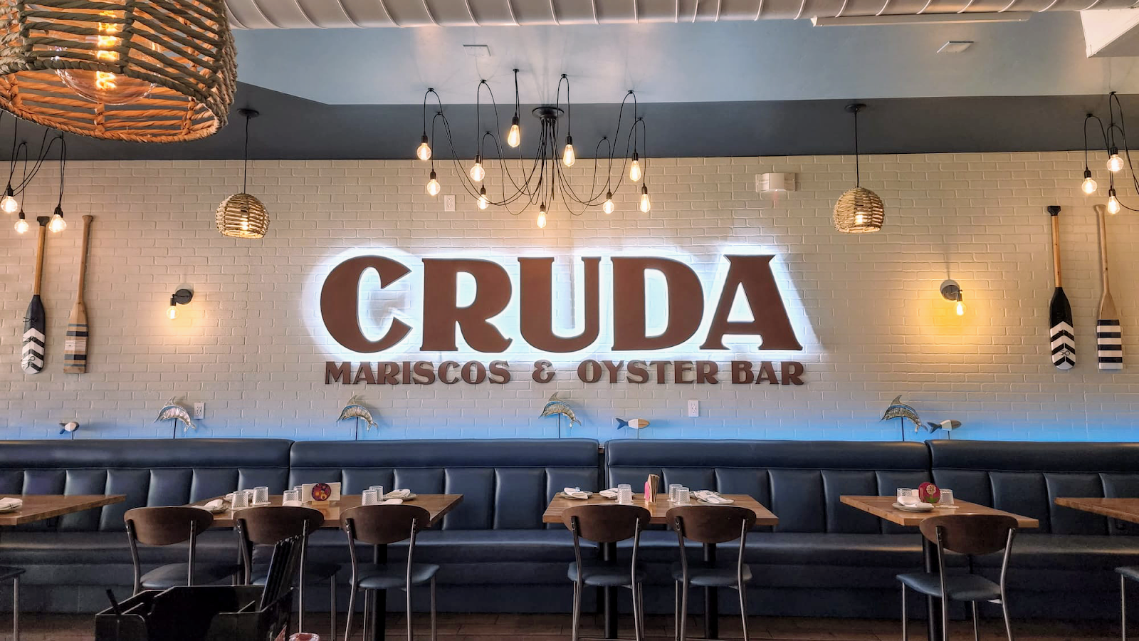 The backlit CRUDA wall sign in rust-red letters on white subway brick, above slate-blue banquettes and hanging Edison bulbs.
