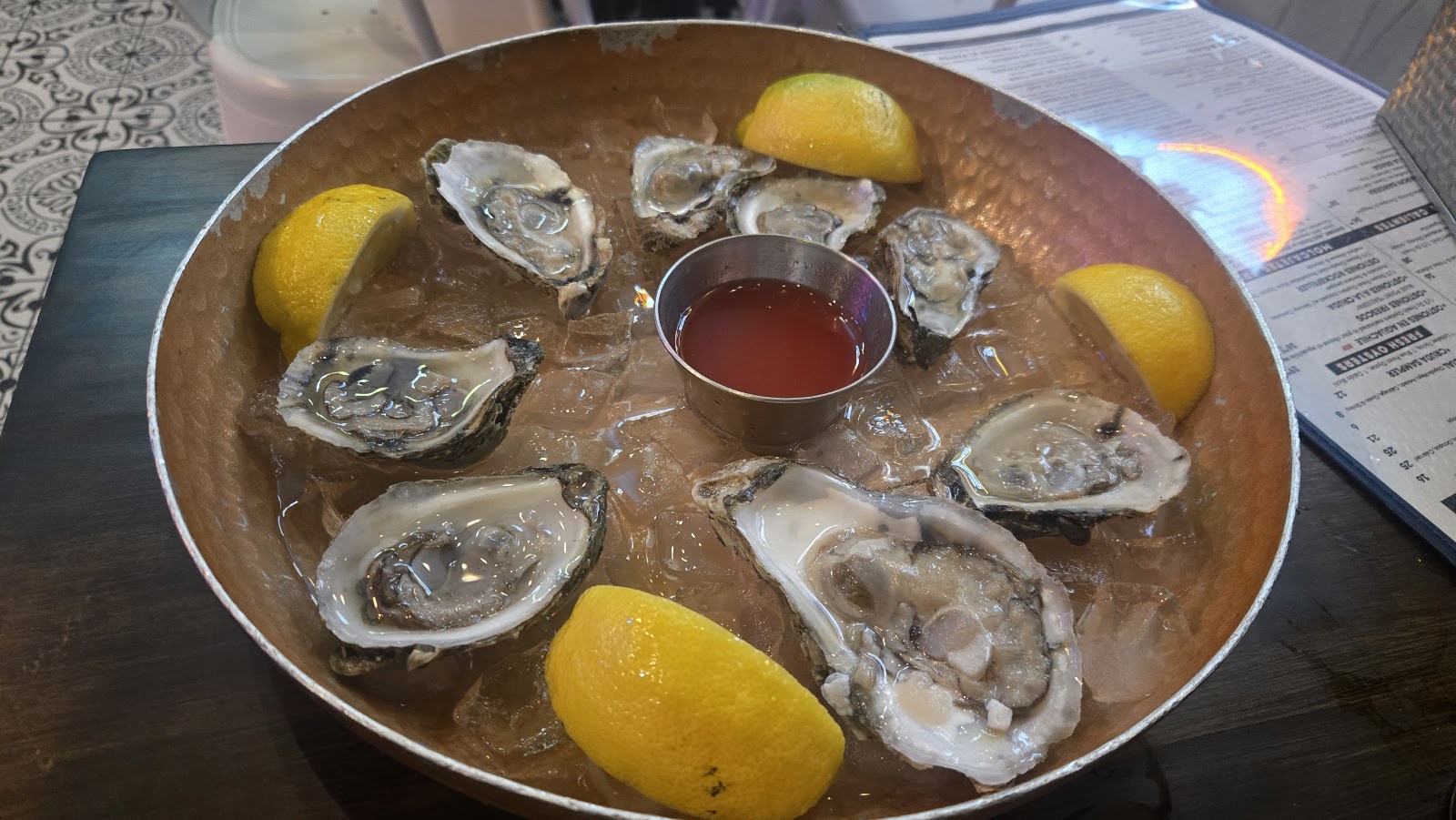 A half-dozen fresh oysters on the half shell over ice, with lemon wedges and a cup of red mignonette in a copper pan.